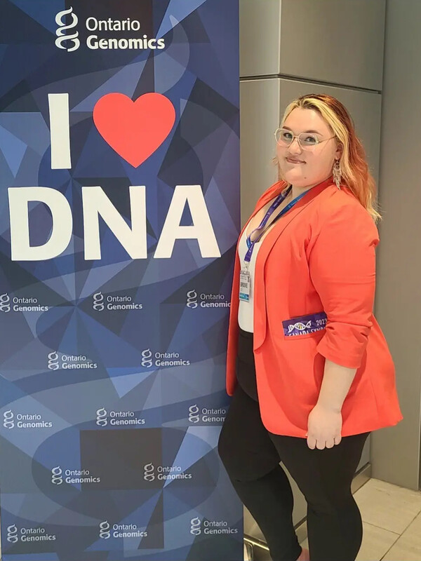 Brooke Coe in a orange blazer posing beside a poster that reads I love DNA