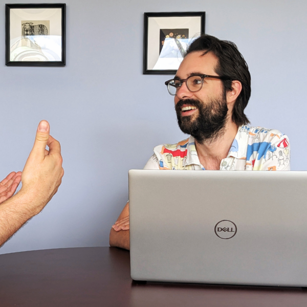 a man smiling at a desk behind his laptop is talking to a friend who hands are only shown